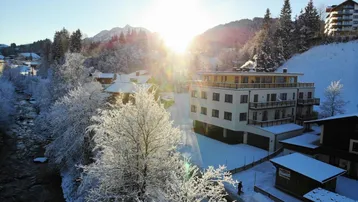 Winterliche Außenansicht eines modernen Gebäudes mit verschneiten Bäumen und Bergen im Hintergrund bei Sonnenaufgang.