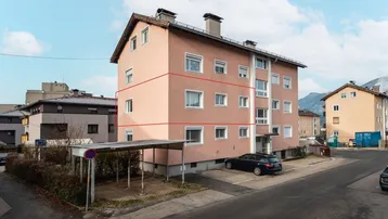 Mehrfamilienhaus mit rosa Fassade, Parkplätzen und Bergblick im Hintergrund.