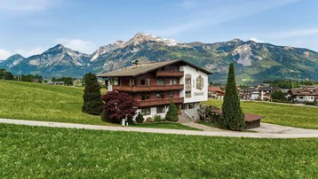 Traditionelles Gebäude im alpinen Stil mit Holzbalkonen, umgeben von grünen Wiesen und majestätischen Bergen.