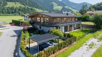 Modernes Haus mit Holzfassade, großzügigen Balkonen, Garten und Pergola mit Whirlpool vor Bergkulisse.