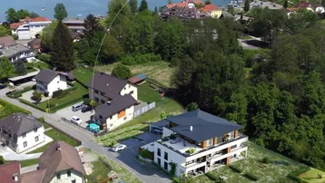 Luftaufnahme eines modernen Neubaus in Seenähe, umgeben von Wohnhäusern und viel Grün.