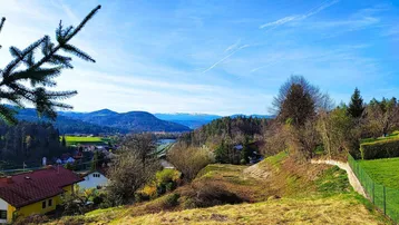 Weitläufiger Blick über die grüne Landschaft mit Häusern und fernen Bergen unter blauem Himmel.