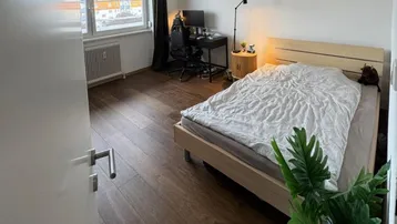Helles Schlafzimmer mit Holzboden, großem Fenster und einem praktischen Arbeitsbereich.