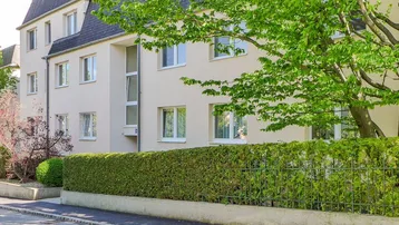 Mehrfamilienhaus mit gepflegter Fassade, umgeben von Grünflächen und Bäumen an einer ruhigen Straße.