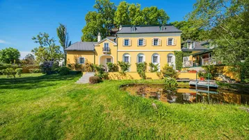 Herrschaftliches Anwesen mit gelber Fassade, großem Garten und idyllischem Teich unter blauem Himmel.