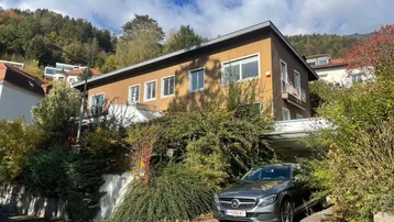 Hausfassade mit üppiger Bepflanzung und einem Carport, eingebettet in eine grüne Umgebung.