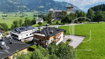 Luftaufnahme eines modernen Gebäudekomplexes in alpiner Landschaft mit Burg und Bergen im Hintergrund.
