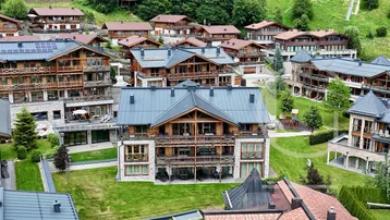 Luftaufnahme eines charmanten Chalet-Dorfes mit modernen Häusern und grünen Gärten vor einer Bergkulisse.
