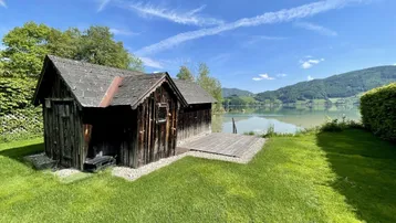 Rustikales Holzhaus am Seeufer mit weitläufigem Rasen und Bergblick unter blauem Himmel.
