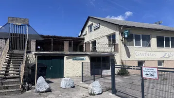 Gebäudeansicht eines Gasthauses mit Außentreppe und Gitterzaun unter blauem Himmel.