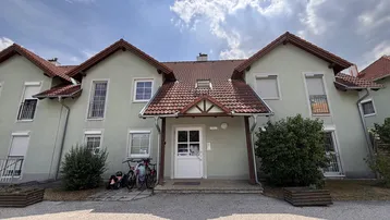 Mehrfamilienhaus mit hellgrüner Fassade, Satteldach und kleinen Balkonen unter bewölktem Himmel.