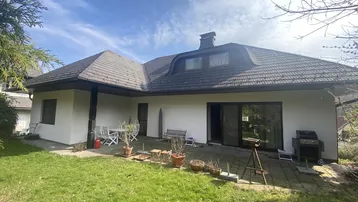 Rückseite des Hauses mit großer gepflasterter Terrasse, Rasenfläche und Gartenmöbeln unter blauem Himmel.