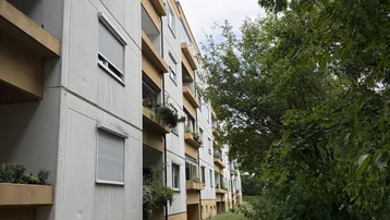 Mehrfamilienhaus mit Balkonen und grüner Umgebung, umgeben von Bäumen und Sträuchern.
