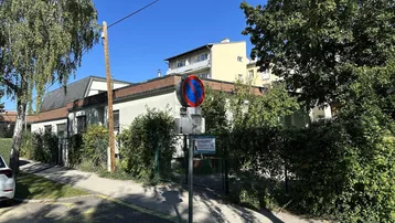 Gebäudeansicht von der Straße mit üppiger Begrünung und einem "Zu Kaufen" Schild.