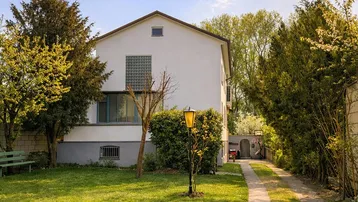 Weißes Einfamilienhaus mit großem Garten, Zufahrt und umgebender Hecke unter blauem Himmel.