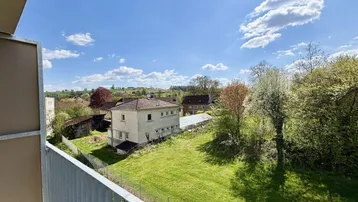 Blick von einem Balkon auf eine ländliche Umgebung mit grünen Wiesen und einem weiteren Gebäude unter blauem Himmel.