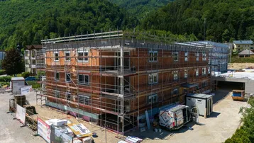 Neubau im Rohbauzustand mit Gerüst vor bewaldeter Bergkulisse unter blauem Himmel.