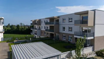 Moderne Wohnanlage mit Balkonen, Gärten und Blick auf grüne Felder unter blauem Himmel.
