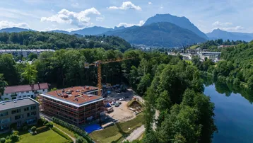 Luftaufnahme einer Baustelle am Seeufer mit Bergen im Hintergrund und einem Kran.