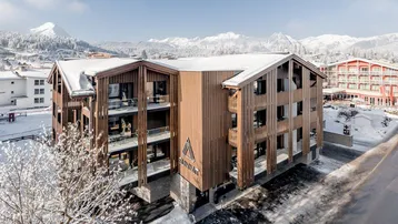 Modernes Gebäude mit Holzfassade und Balkonen in verschneiter Berglandschaft unter blauem Himmel.