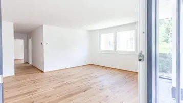 Heller, leerer Wohnraum mit Holzboden und Zugang zu einem Balkon.