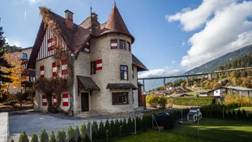 Historisches Gebäude mit Turm und roten Fensterläden, umgeben von einem Garten und Bergblick.