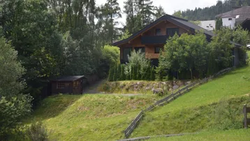 Modernes Holzhaus im Chalet-Stil auf einem grünen Hügel, umgeben von Bäumen und Natur, mit einem kleinen Gartenhaus.