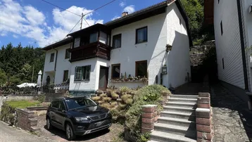 Weißes Wohnhaus mit Balkon und Außentreppe, umgeben von grüner Vegetation unter blauem Himmel.