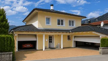 Gelbes Einfamilienhaus mit Doppelgarage und gepflasterter Einfahrt unter blauem Himmel.