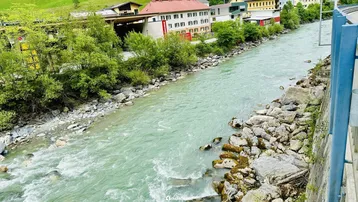 Flusslandschaft mit felsigen Ufern, umgeben von Gebäuden und grünen Hügeln unter bewölktem Himmel.