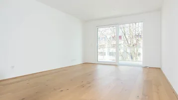 Helles, leeres Zimmer mit Holzboden und Zugang zu einem Balkon oder einer Terrasse.