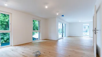 Helles, leeres Wohnzimmer mit neuem Parkettboden und großen Fenstern, die viel Tageslicht hereinlassen.