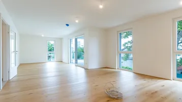 Helles, leeres Wohnzimmer mit neuem Parkettboden und großen Fenstern, die viel Tageslicht hereinlassen.