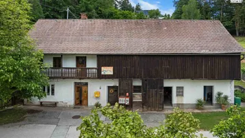 Traditionelles Bauernhaus mit weißer Fassade, Holzbalkon und großem Scheunentor, umgeben von grünen Bäumen.