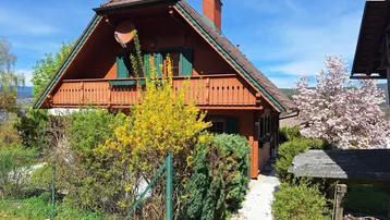 Charmantes Holzhaus mit Balkon, grünen Fensterläden und blühenden Sträuchern unter blauem Himmel.