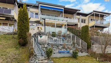 Mehrfamilienhaus mit terrassiertem Gartenbereich, Außentreppe und Balkonen unter blauem Himmel.