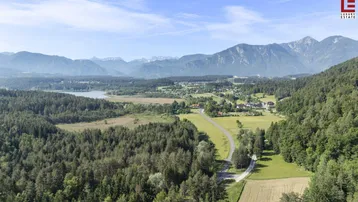 Weitläufige Landschaftsaufnahme mit Wäldern, Feldern, einem kleinen See und majestätischen Bergen im Hintergrund.