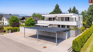Modernes Wohngebäude mit Carport und Balkonen in grüner Umgebung unter blauem Himmel.