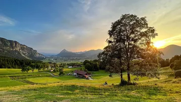 Weitläufige Landschaft mit grünen Wiesen, majestätischen Bergen und einem malerischen Sonnenuntergang.