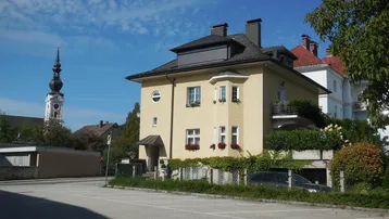 Historisches Gebäude mit gelber Fassade und Kirchturm im Hintergrund unter blauem Himmel.