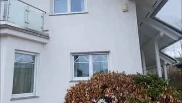Weiße Hausfassade mit Balkon und Satteldach, umgeben von winterlicher Vegetation.
