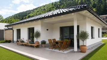 Modernes Einfamilienhaus mit großer Sonnenterrasse, Gartenmöbeln und Blick auf die bewaldeten Berge.