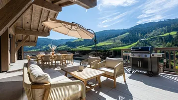 Geräumige Terrasse mit bequemen Sitzgelegenheiten, Grillbereich und herrlichem Bergblick unter blauem Himmel.