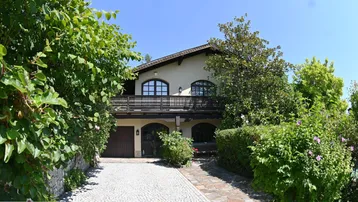 Charmantes Haus mit gepflasterter Auffahrt, üppigem Grün und einem Balkon im Obergeschoss.