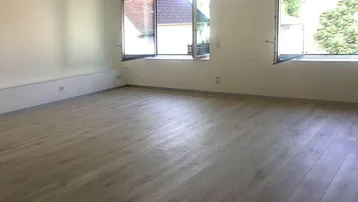 Heller, leerer Raum mit großen Fenstern und modernem Laminatboden, ideal als Wohnzimmer.