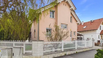 Rosa gestrichenes Einfamilienhaus mit weißem Zaun und gepflegtem Vorgarten unter blauem Himmel.