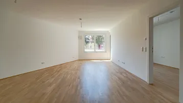 Helles, leeres Wohnzimmer mit Parkettboden und einem Fenster mit Blick ins Grüne.