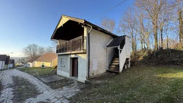 Altes Landhaus mit Balkon und Außentreppe in ländlicher Umgebung unter blauem Himmel.
