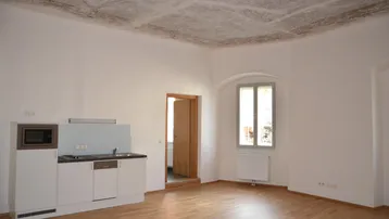 Helles Wohnzimmer mit integrierter Küchenzeile, Parkettboden und historischer Deckenverzierung.