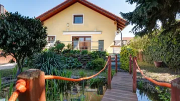 Charmantes Haus mit gepflegtem Garten, Teich und kleiner Holzbrücke unter blauem Himmel.